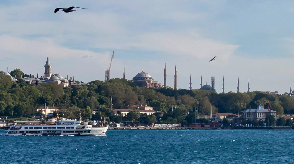 Vista de Istambul a partir do Bósforo, com barco no primeiro plano, mar azul, aves no céu e a silhueta histórica da cidade ao fundo, na Turquia.