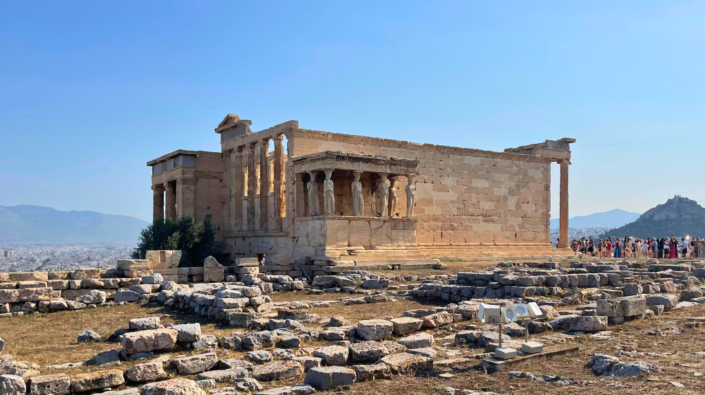 Erecteion na Acrópole de Atenas, na Grécia, com ruínas de pedra, visitantes ao fundo e céu azul.