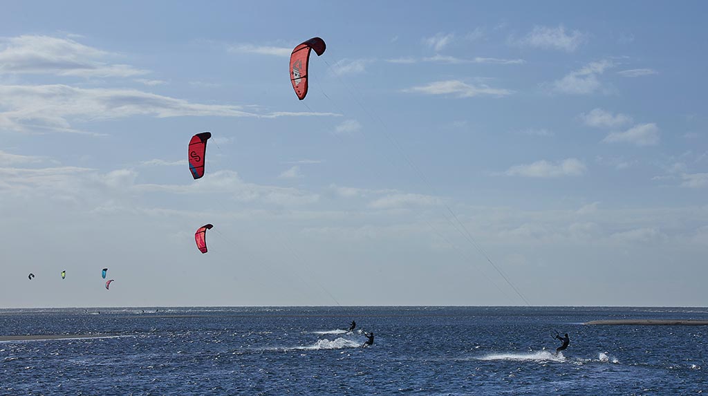Kitesurfistas navegando em mar raso com vento forte, dunas ao fundo e céu limpo.