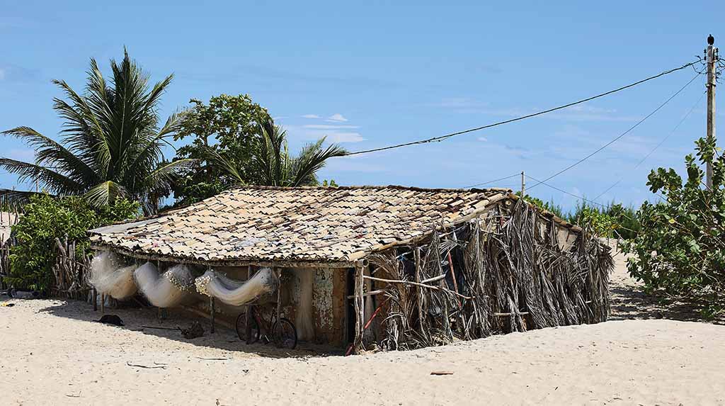 Casa de taipa com cobertura simples, cercada por areia e vegetação nativa da Praia Nova.