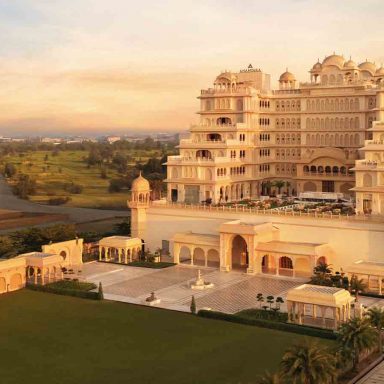 Fachada palaciana do Anantara Jewel Bagh Jaipur ao pôr do sol, com cúpulas e jharokas em pedra dourada, pátios simétricos e jardins amplos nos arredores de Jaipur, Índia.