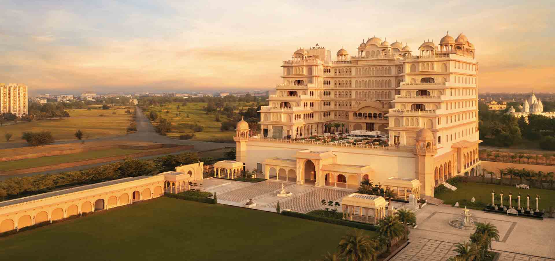 Fachada palaciana do Anantara Jewel Bagh Jaipur ao pôr do sol, com cúpulas e jharokas em pedra dourada, pátios simétricos e jardins amplos nos arredores de Jaipur, Índia.