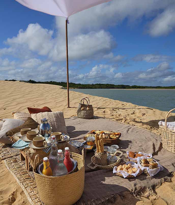 Mesa posta sobre as dunas, com frutas e pães, ao lado do rio, sob guarda-sol.