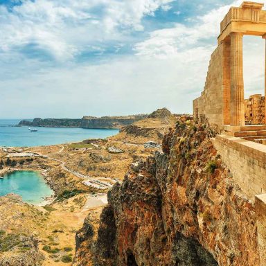 Acrópole de Líndos (Rodes): ruínas dóricas e muralhas medievais no topo do penhasco, vila caiada e baías azul-turquesa do mar Egeu ao fundo.
