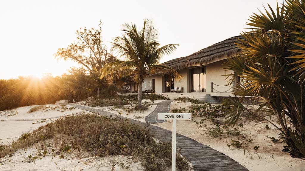 Caminho de madeira levando à entrada da residência privativa 'Cove One' no Kisawa Sanctuary, cercada por vegetação nativa ao pôr do sol.