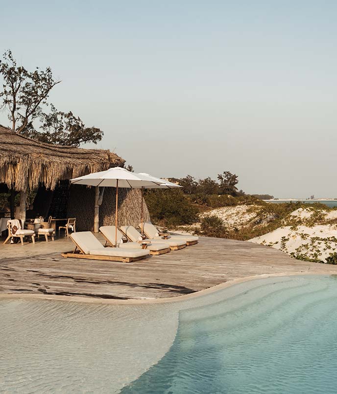 Área de lazer luxuosa do Kisawa Sanctuary com piscina de borda curva, espreguiçadeiras e vista para as dunas de areia branca e o mar.