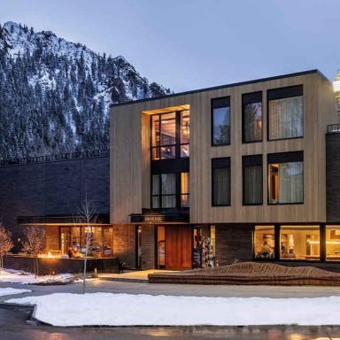Fachada do Mollie Aspen, hotel boutique em Aspen, Colorado, com neve na rua e montanhas ao fundo ao entardecer