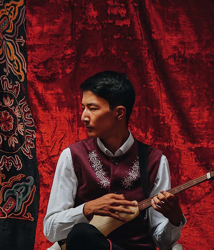 Músico tradicional quirguiz tocando komuz, instrumento de cordas típico da cultura nômade, em cenário decorado com tecido bordado