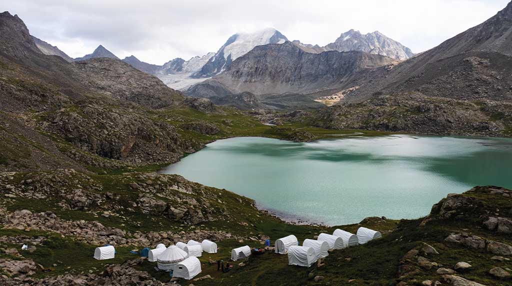Acampamento de yurts brancas às margens do lago de montanha Ala-Kul, cercado por picos rochosos e neve eterna, Quirguistão