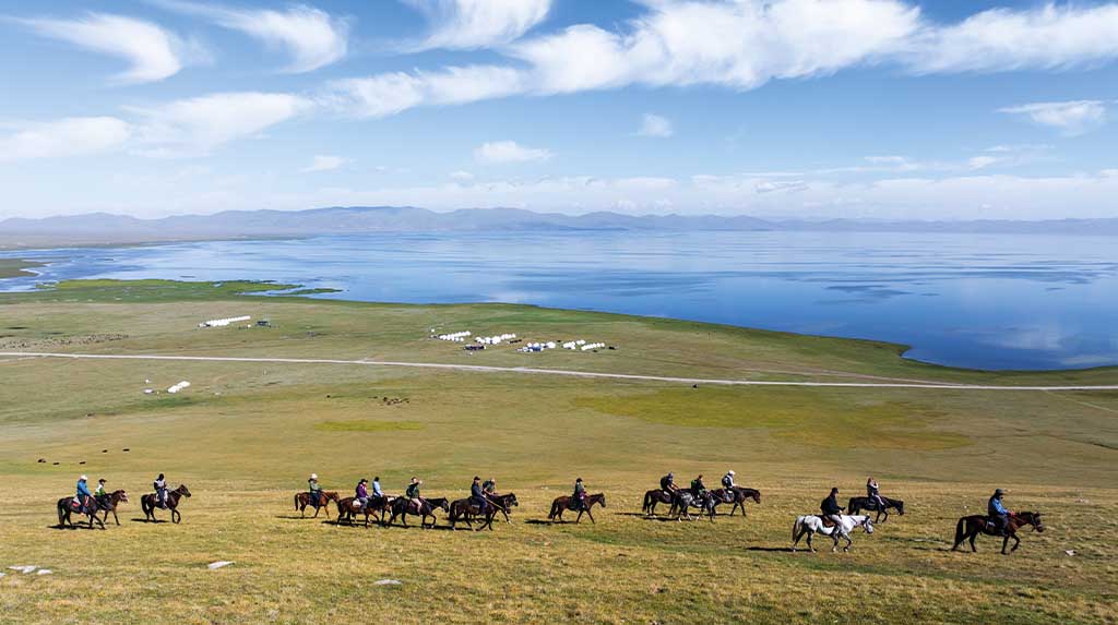 Grupo de viajantes a cavalo percorrendo as margens do Lago Song-Kul, no Quirguistão, com planícies verdes e yurts ao fundo