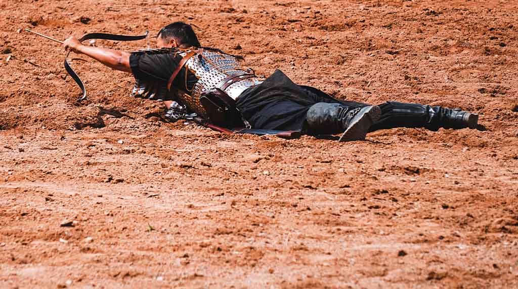 Competidor quirguiz de arco e flecha deitado no chão durante apresentação tradicional do Salburun Festival, com traje histórico nômade