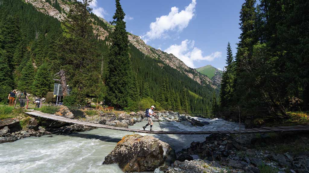 Trilha em ponte de madeira sobre rio glacial no Ala-Archa National Park, Quirguistão, com montanhas e florestas densas ao redor