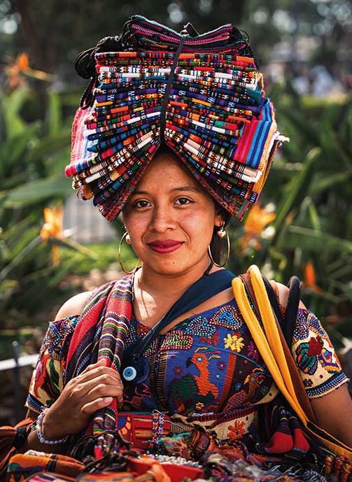 Vendedora guatemalteca maia sorridente em Antígua, Guatemala, vestindo um huipil tradicional ricamente bordado em tons de azul e roxo. Ela equilibra na cabeça uma alta pilha de tecidos artesanais de cores vibrantes, contrastando com o fundo de folhagens verdes e flores vermelhas desfocadas.