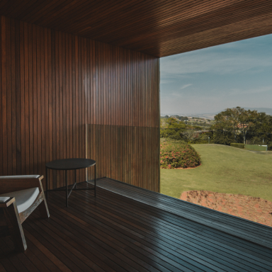 Varanda de madeira no Hotel Fasano Boa Vista, em Porto Feliz (SP), com poltrona e mesa, enquadrando a vista verde da Fazenda Boa Vista e colinas ao fundo.