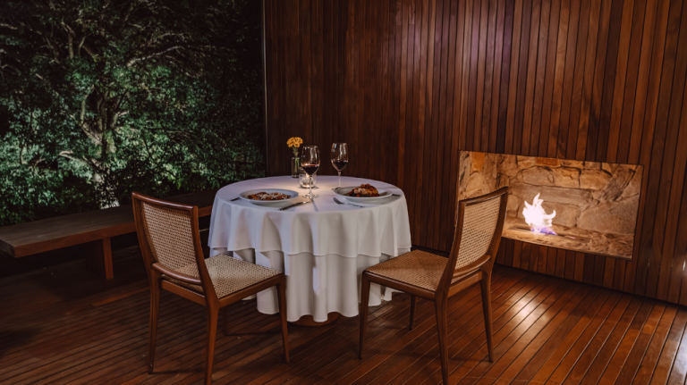 Mesa para dois com toalha branca e taças de vinho no restaurante do Hotel Fasano Boa Vista, em Porto Feliz (SP), com cadeiras de palhinha, madeira aparente e lareira acesa ao lado Mesa para dois com toalha branca e taças de vinho no restaurante do Hotel Fasano Boa Vista, em Porto Feliz (SP)