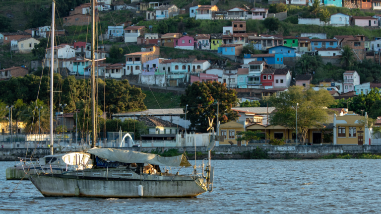 Barco à vela em rio largo diante de casario colorido em encosta na cidade de Cachoeira, no Recôncavo Baiano, com luz suave de fim de tarde
