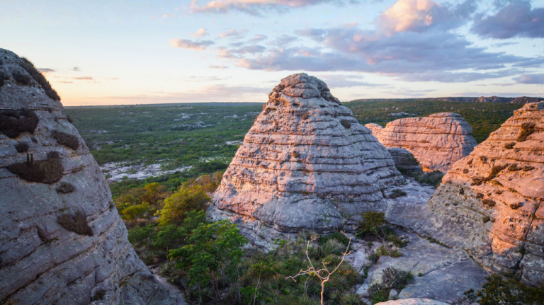 Formações rochosas monumentais na caatinga do Parque Nacional do Catimbau ao pôr do sol, com vegetação baixa e céu em tons pastel
