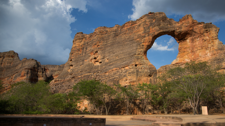 Arco natural de arenito conhecido como Pedra Furada no Parque Nacional da Serra da Capivara, Piauí, com vegetação de caatinga e nuvens densas ao fundo.
