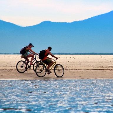 Ciclistas pedalam na faixa de areia de uma praia ampla no litoral do Paraná, com mar calmo em primeiro plano e montanhas azuladas ao fundo, Brasil.