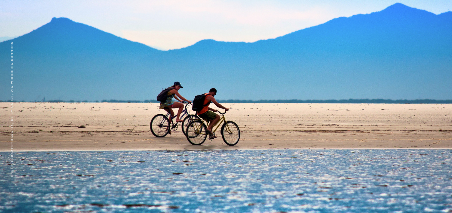 Ciclistas pedalam na faixa de areia de uma praia ampla no litoral do Paraná, com mar calmo em primeiro plano e montanhas azuladas ao fundo, Brasil.