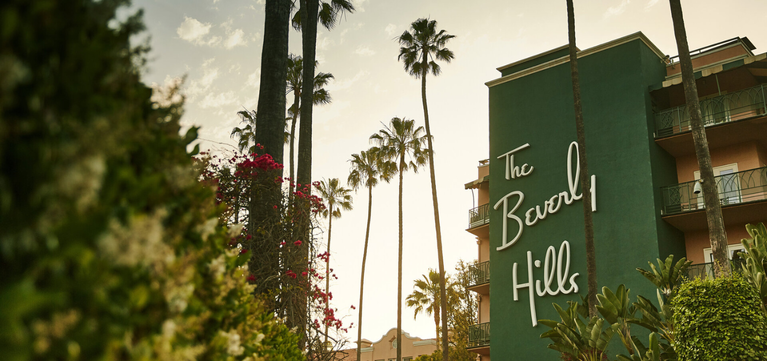 Letreiro verde do The Beverly Hills Hotel entre palmeiras ao entardecer, em Beverly Hills, Los Angeles — ponto de partida do tour Building Dreams in Beverly Hills inspirado em Paul R. Williams.”