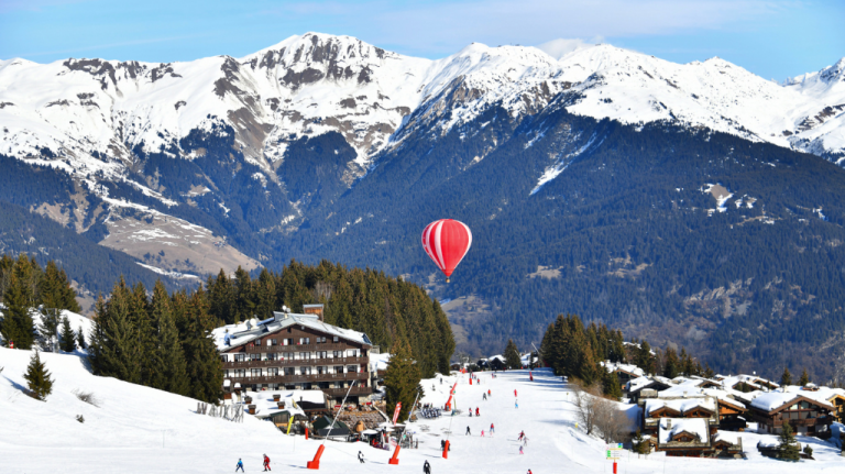 Pista de esqui em Courchevel, nos Alpes franceses, com chalés de montanha, pinheiros nevados, esquiadores e um balão de ar quente diante dos picos brancos Pista de esqui em Courchevel, nos Alpes franceses, com chalés de montanha