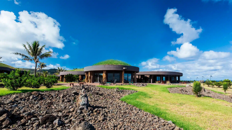Fachada do Nayara Hangaroa, hotel em Rapa Nui, na Ilha de Páscoa, Chile, com arquitetura de linhas circulares, telhado verde, jardins abertos e paisagismo sobre terreno vulcânico sob céu azul intenso; imagem ideal para conteúdos sobre hotel de luxo no Chile, hospedagem em Rapa Nui, Ilha de Páscoa, arquitetura integrada à natureza, viagem ao Pacífico Sul e turismo sustentável.