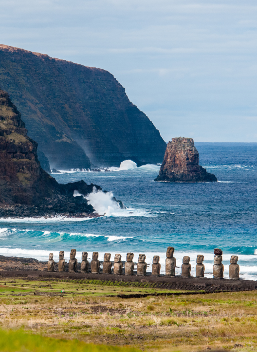 Paisagem costeira de Rapa Nui, na Ilha de Páscoa, a cerca de 3.700 km de Santiago, com fileira de moais voltados para o interior da ilha, ondas do oceano Pacífico, falésias vulcânicas e um rochedo isolado ao fundo; imagem marcante para conteúdos sobre Ilha de Páscoa, Rapa Nui, moais, patrimônio da UNESCO, viagem ao Chile, arqueologia e paisagem do Pacífico Sul.