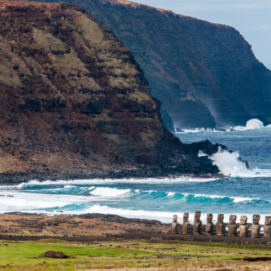 Paisagem costeira de Rapa Nui, na Ilha de Páscoa, com uma fileira de moais sobre uma plataforma de pedra diante do oceano Pacífico, ondas brancas, mar em tons de azul-turquesa e falésias vulcânicas monumentais ao fundo. A cena combina arqueologia, natureza dramática e patrimônio cultural em um dos cenários mais emblemáticos do Chile, ideal para conteúdos sobre Ilha de Páscoa, Rapa Nui, moais, viagem ao Chile, sítios arqueológicos e paisagens do Pacífico Sul.