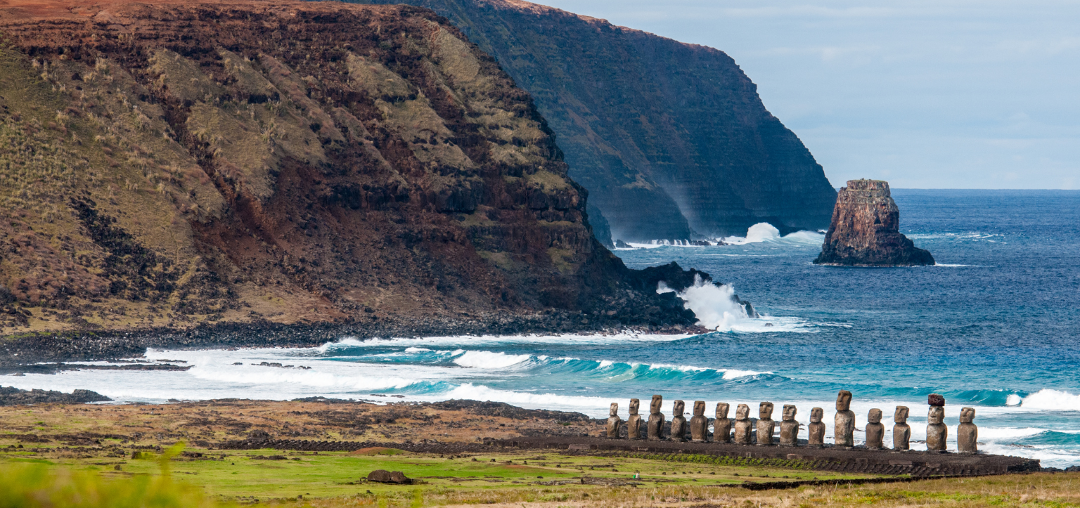 Paisagem costeira de Rapa Nui, na Ilha de Páscoa, com uma fileira de moais sobre uma plataforma de pedra diante do oceano Pacífico, ondas brancas, mar em tons de azul-turquesa e falésias vulcânicas monumentais ao fundo. A cena combina arqueologia, natureza dramática e patrimônio cultural em um dos cenários mais emblemáticos do Chile, ideal para conteúdos sobre Ilha de Páscoa, Rapa Nui, moais, viagem ao Chile, sítios arqueológicos e paisagens do Pacífico Sul.