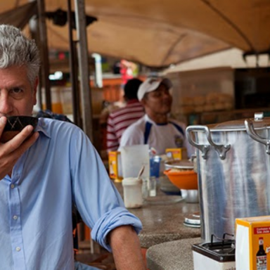 Anthony Bourdain sentado no mercado Ver-o-Peso, em Belém do Pará, tomando tacacá em uma mesa simples, com barracas e recipientes de comida ao fundo.