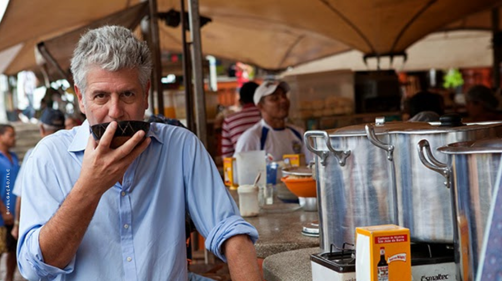 Anthony Bourdain sentado no mercado Ver-o-Peso, em Belém do Pará, tomando tacacá em uma mesa simples, com barracas e recipientes de comida ao fundo.