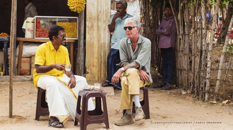 Anthony Bourdain sentado ao ar livre com outra pessoa, em cenário de rua, durante uma conversa de viagem. A imagem sugere escuta, encontro, comida como experiência cultural e o método de Anthony Bourdain para entender destinos pela gastronomia Anthony Bourdain sentado ao ar livre, em cenário de rua, durante uma conversa de viagem