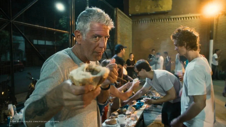 Foto mostra Anthony Bourdain em uma cozinha movimentada, cercado por cozinheiros e utensílios, durante uma gravação gastronômica. A cena destaca o contato direto com quem cozinha e reforça temas como jornalismo gastronômico, cultura local, viagem pela comida e bastidores da cozinha O melhor mapa de Bourdain sempre foi humano