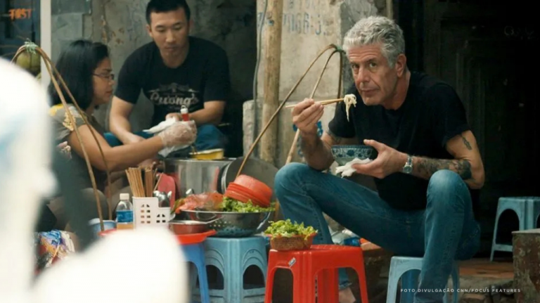 Anthony Bourdain com outra pessoa em uma mesa baixa de comida de rua em Hanói, capital do Vietnã, no norte do país, com pratos e bebidas sobre a mesa. A imagem remete ao bún chả, à comida de rua vietnamita, aos bancos baixos, aos caldos, às ervas frescas e à viagem gastronômica no Vietnã Anthony Bourdain em uma mesa baixa de comida de rua em Hanói, Vietnã