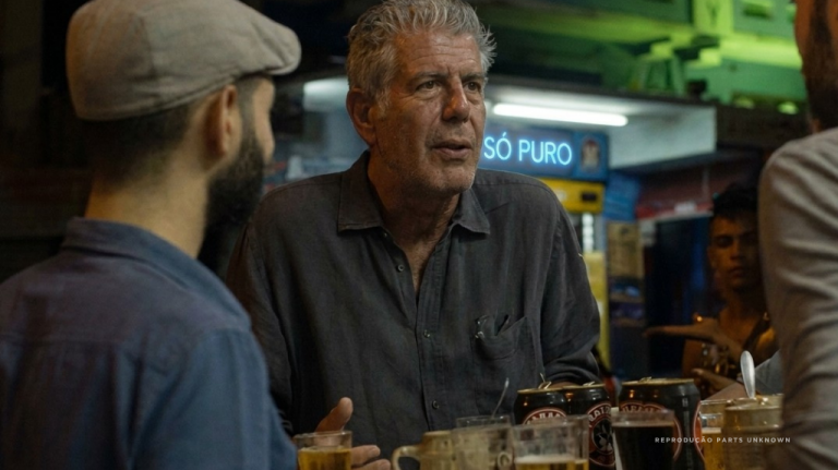 Foto mostra Anthony Bourdain em uma conversa informal dentro de um mercado ou boteco em Belo Horizonte, capital de Minas Gerais, na Região Sudeste do Brasil. A imagem remete à cozinha mineira, aos balcões populares, ao Mercado Central, aos botecos de BH e à viagem gastronômica pelo Brasil. Anthony Bourdain conversando em um balcão em BH
