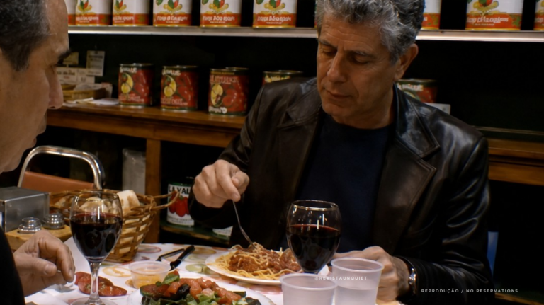 Anthony Bourdain sentado em uma deli de Nova York, no Nordeste dos Estados Unidos, diante de comida servida em mesa simples, com ambiente casual ao fundo. A imagem remete às delis nova-iorquinas, ao pastrami, ao pão de centeio, à imigração judaica e à gastronomia histórica de bairro Anthony Bourdain sentado em uma deli de Nova York, diante de comida servida em mesa simples, com ambiente casual ao fundo. A imagem remete às delis nova-iorquinas, ao pastrami, ao pão de centeio, à imigração judaica e à gastronomia histórica de bairro