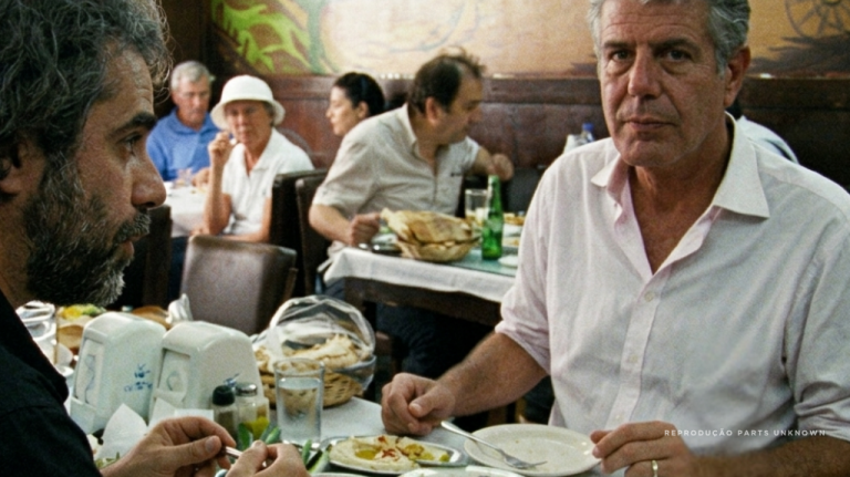 Anthony Bourdain diante de uma mesa compartilhada com pratos, pães e pessoas ao redor, em contexto associado a Beirute, capital do Líbano. A imagem evoca mezze, hospitalidade libanesa, memória cultural, comida como reconstrução e viagem gastronômica no Oriente Médio. Anthony Bourdain diante de uma mesa compartilhada com pratos, pães e pessoas ao redor