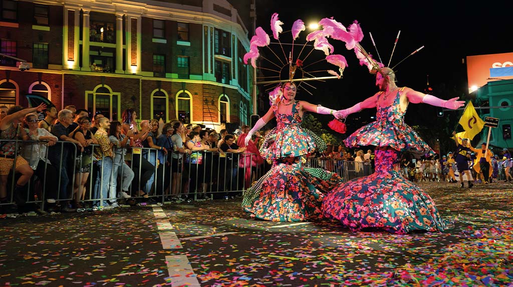 Desfile noturno do Orgulho LGBTQIA+ com performer fantasiado e público nas ruas, em celebração de cultura queer e diversidade.