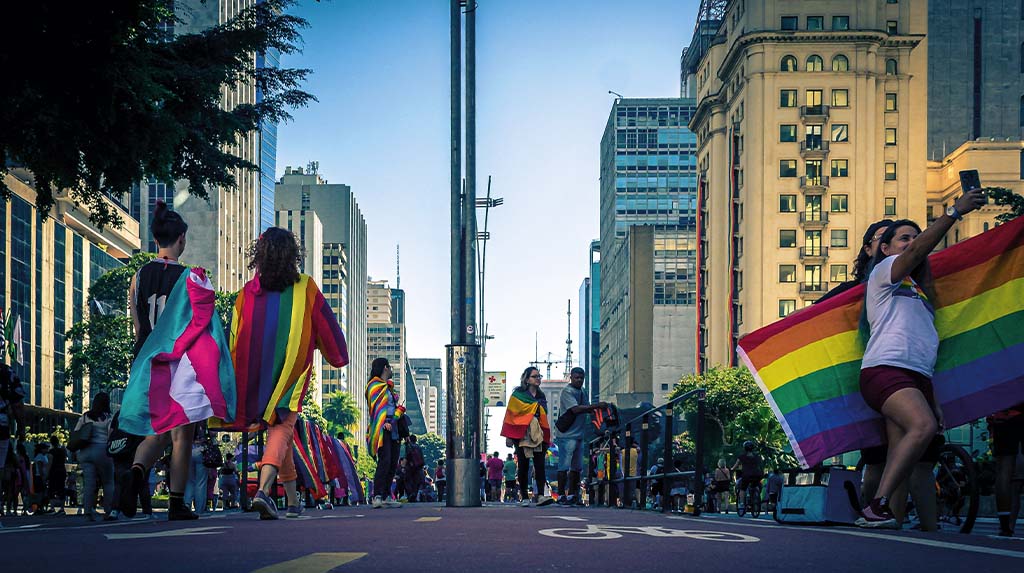 Bandeiras do arco-íris ocupam avenida durante celebração do Orgulho LGBTQIA+, em cena urbana associada à visibilidade e à luta por direitos.