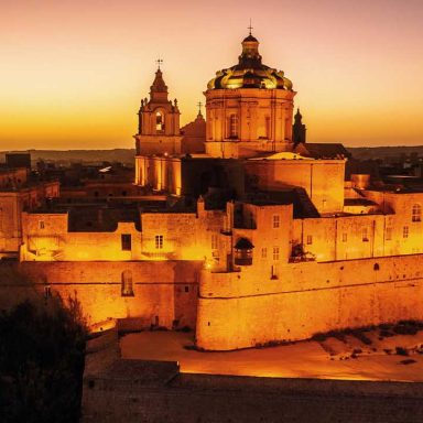 Vista aérea de Mdina, cidade murada histórica na Ilha de Malta, com a Catedral de São Paulo iluminada ao entardecer, muralhas medievais e arquitetura barroca no centro do Mediterrâneo.