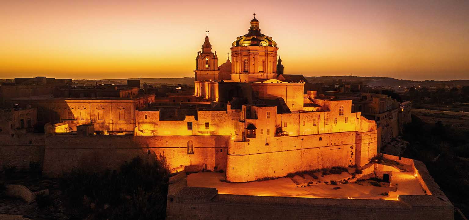 Vista aérea de Mdina, cidade murada histórica na Ilha de Malta, com a Catedral de São Paulo iluminada ao entardecer, muralhas medievais e arquitetura barroca no centro do Mediterrâneo.