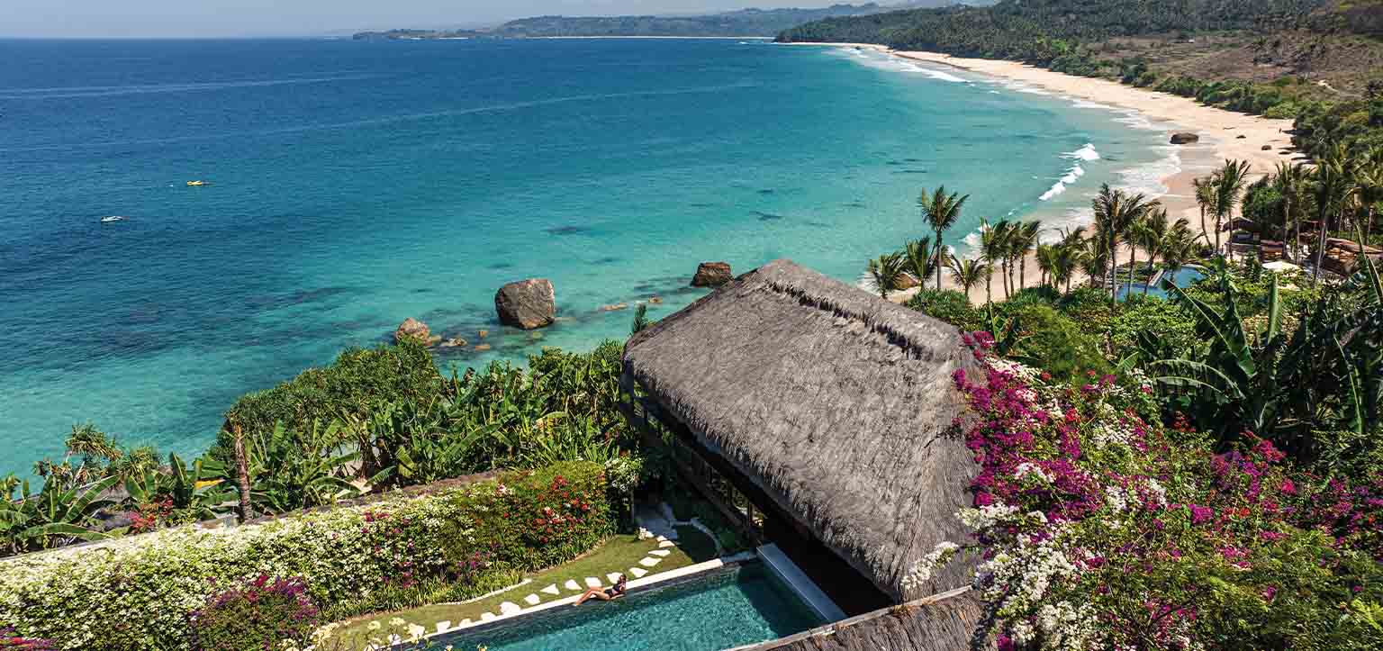 Vista aérea do Nihi Sumba, hotel de luxo na Ilha de Sumba, no leste da Indonésia, com villa de telhado tradicional, piscina privativa, jardins tropicais, praia preservada e mar azul-turquesa do Oceano Índico.